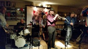 'Hershey and the Hotshots' playing for the first time at Farnborough Jazz Club on 4th December 2015. (LtoR) Paul Taylor (trombone), (Keith Grant has sit-in on drums), Roger Curphey (double bass), Peter Leonard (trumpet&vocs), Tim Huskisson (clarinet&alto sax) and (out of view) Andrew Clancy (piano) and drummer Graham Caulicott dances with Jill. Photo by Peter Marr.