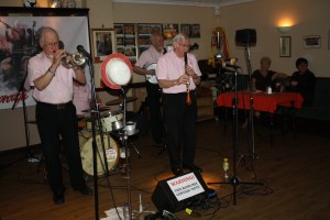 Dave Stradwick (trumpet), Pete Lay (drums), Phil Durell (banjo) and Bernard Stutt (clarinet) in Sussex Jazz Kings at Farnborough Jazz Club on 29th April 2016. Seated right - at table is band organiser Lesley Stradwick with Heather Lay. Photo by Mike Witt.